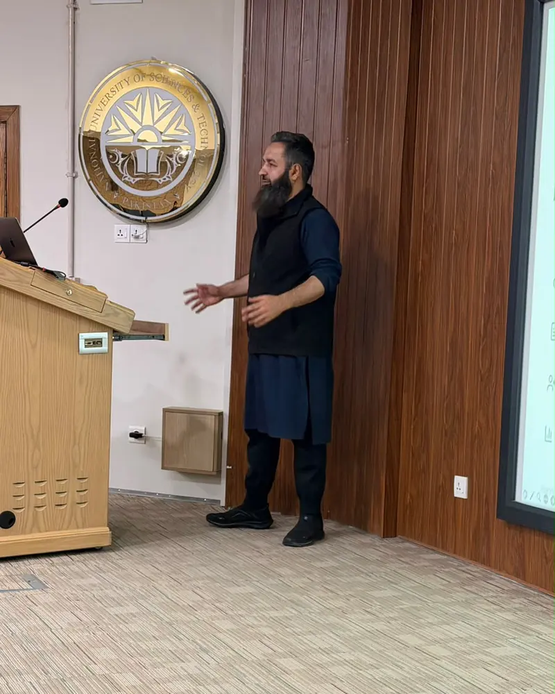 Umar Muhammad Sharif speaking beside a podium in a university lecture room, with a crest on the wall
